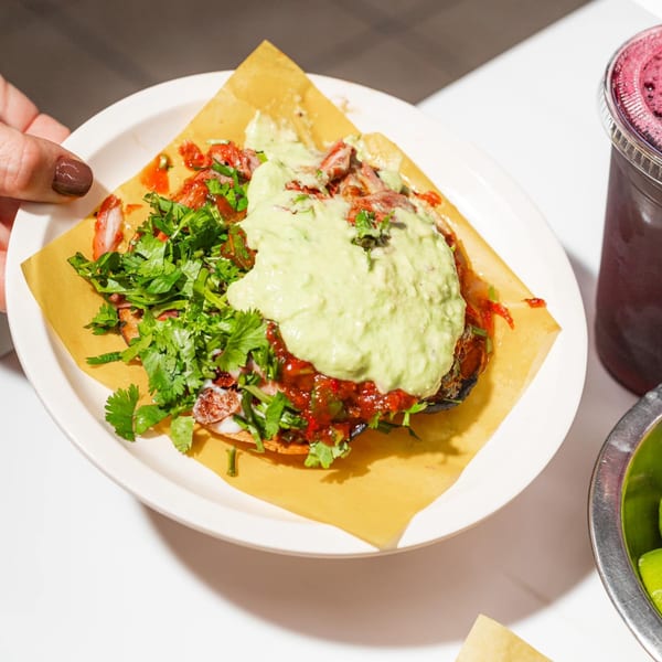 A hand holds a plate with a Tacos El Franc taco topped with meat, salsa, guacamole, and cilantro.