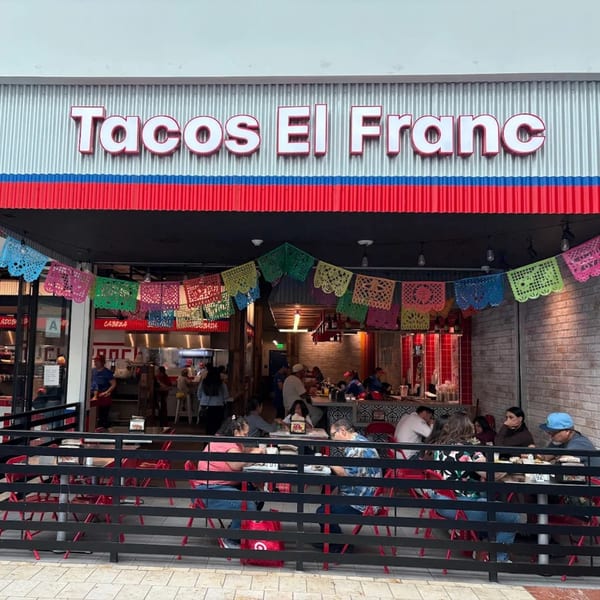 Tacos El Franc restaurant exterior with outdoor seating, colorful papel picado, and patrons dining.