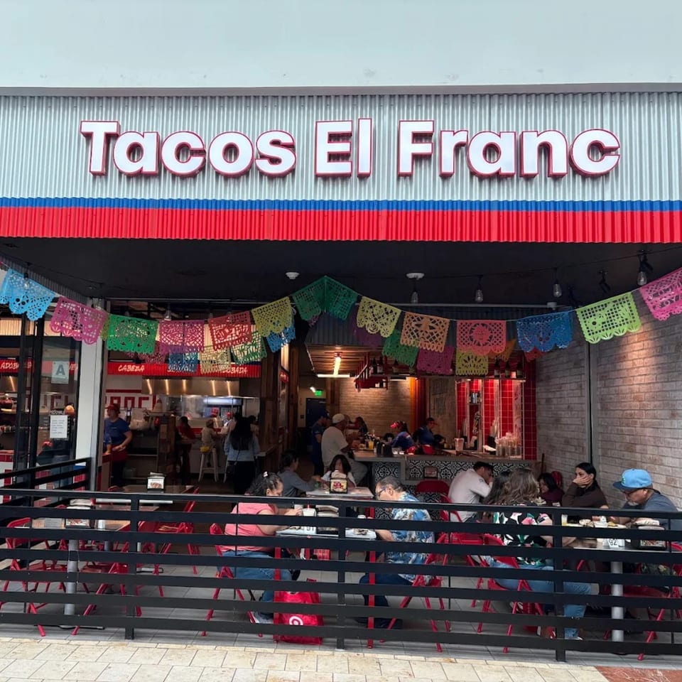 Tacos El Franc restaurant exterior with outdoor seating, colorful papel picado, and patrons dining.