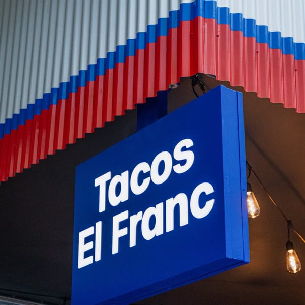 Blue and white Tacos El Franc sign hangs below a red, white, and blue corrugated awning.