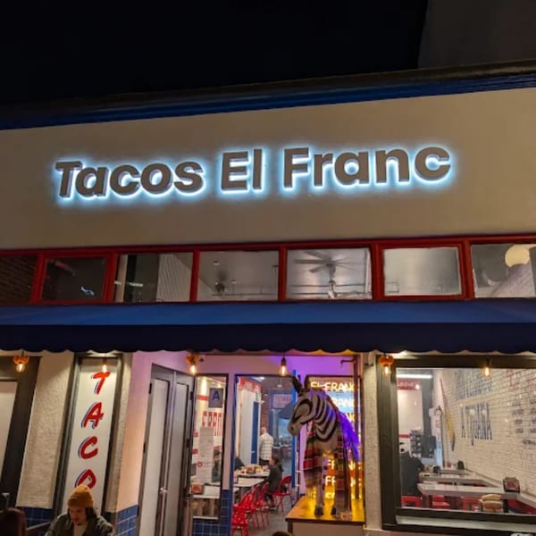 Exterior of Tacos El Franc at night, with a glowing sign and a zebra statue in the window.