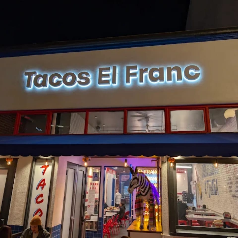 Exterior of Tacos El Franc at night, with a glowing sign and a zebra statue in the window.