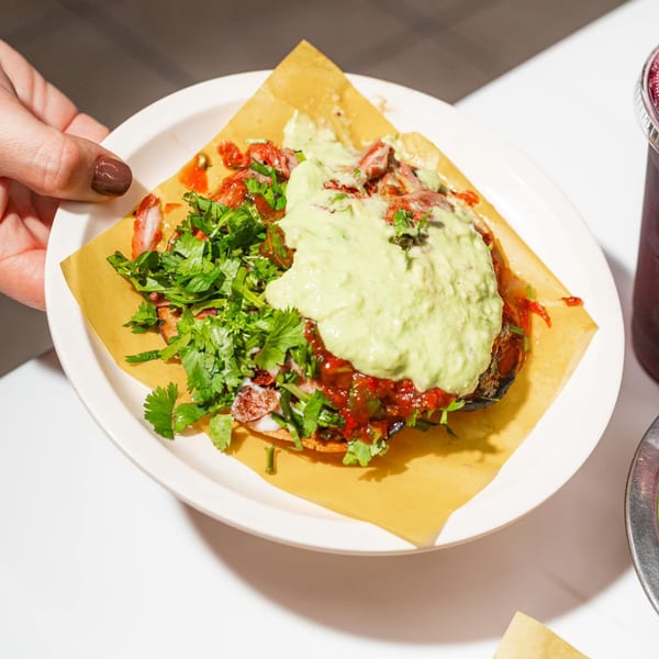 A hand holds a plate with a Tacos El Franc taco, topped with meat, salsa, guacamole, and cilantro.