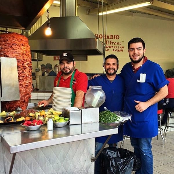 Three Tacos El Franc employees smiling behind a counter with a large al pastor trompo.