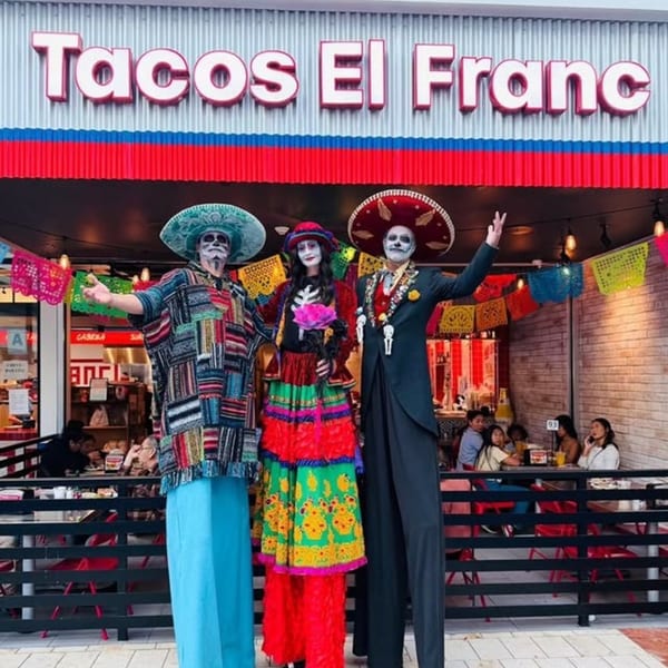 Three people in colorful Day of the Dead costumes stand outside Tacos El Franc restaurant.