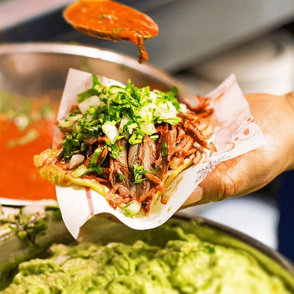 A hand holding a Tacos El Franc taco with meat, cilantro, onions, and red sauce being drizzled.