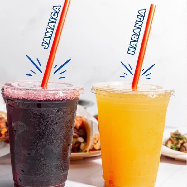 Two plastic cups of Tacos El Franc aguas frescas, Jamaica and Naranja, with tacos in the background.