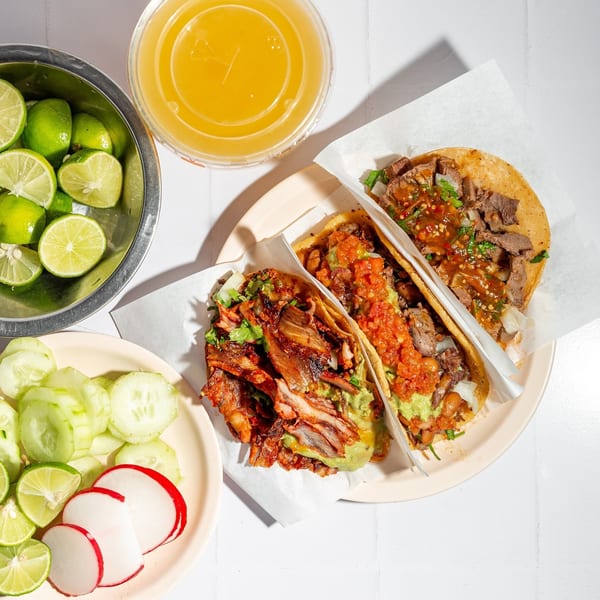 Two Tacos El Franc tacos with various toppings, limes, cucumbers, radishes, and a drink.