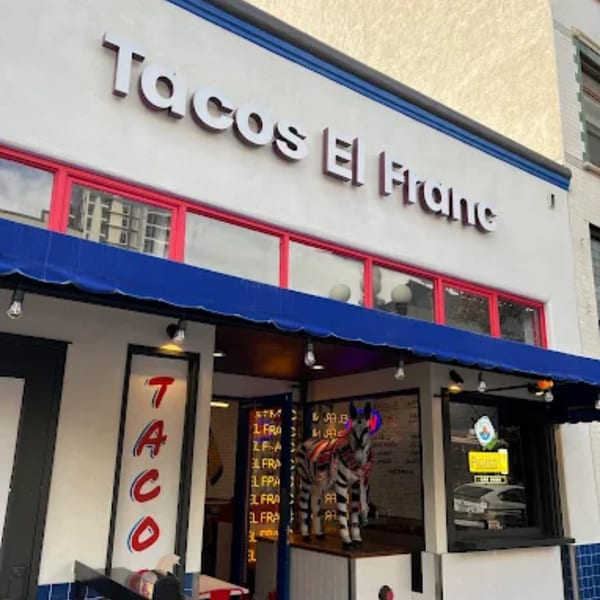 The exterior of Tacos El Franc restaurant with a blue awning and a zebra statue in the window.