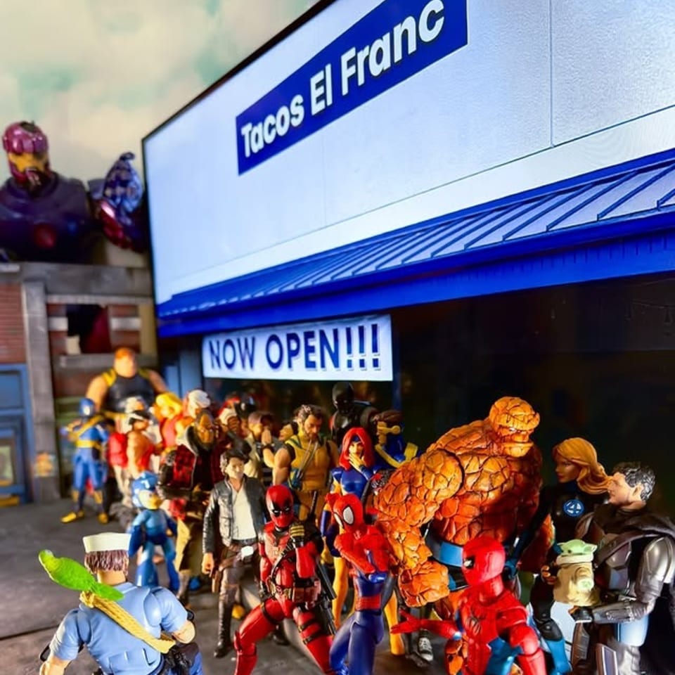 Superhero action figures line up outside Tacos El Franc, with a "NOW OPEN!!!" banner displayed.