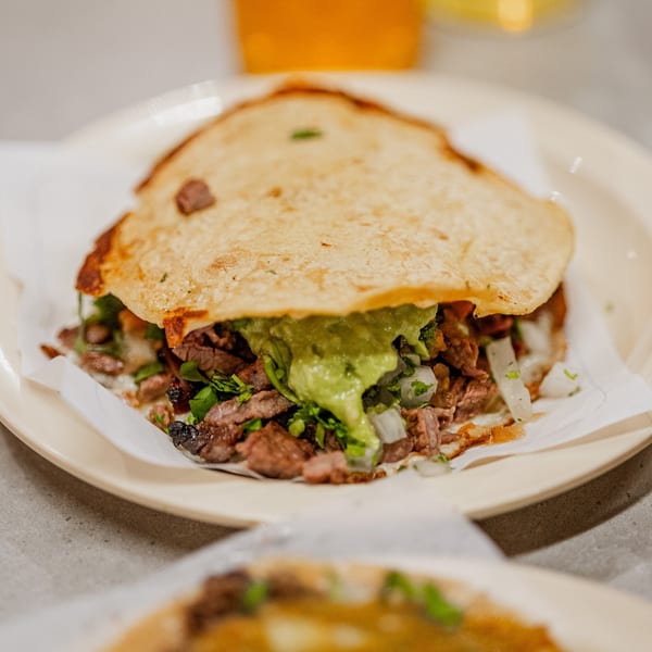 A Tacos El Franc taco with carne asada, guacamole, onions, and cilantro on a white plate.
