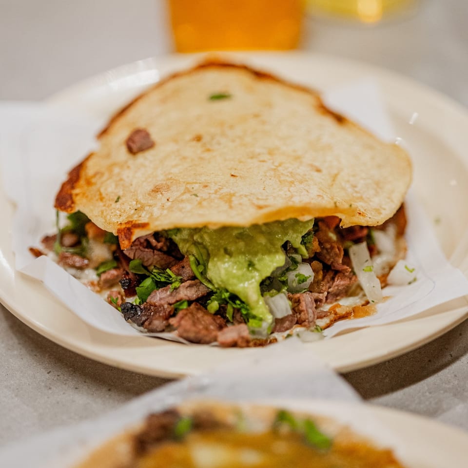 A Tacos El Franc taco with carne asada, guacamole, onions, and cilantro on a white plate.