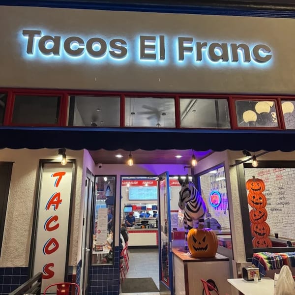 The exterior of Tacos El Franc restaurant at night, with a glowing sign and Halloween decorations.