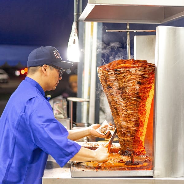Chef carving steaming al pastor from a vertical rotisserie at Tacos El Franc.