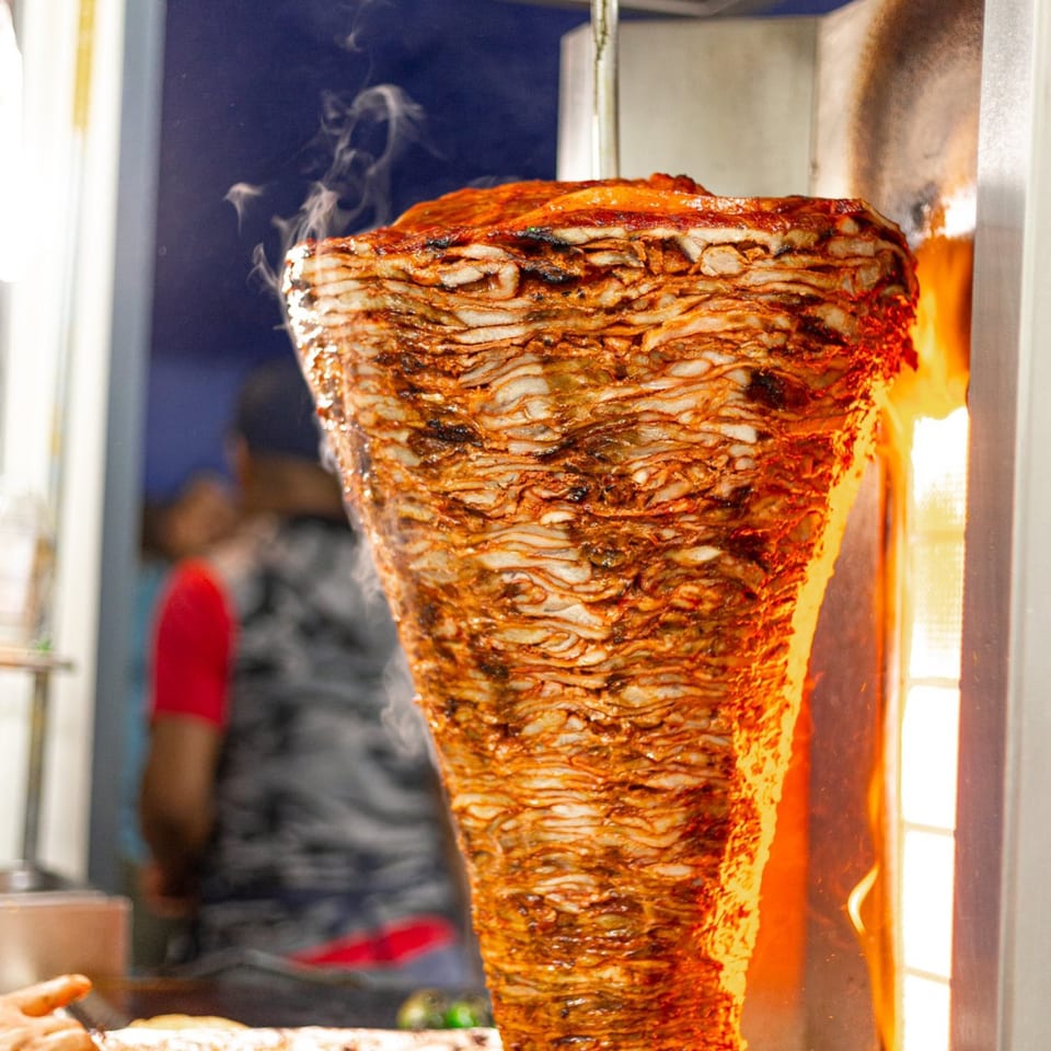 Vertical spit of sizzling al pastor meat cooking over an open flame at Tacos El Franc.