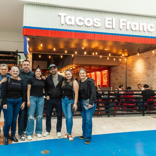 Six people smiling in front of the Tacos El Franc restaurant entrance with string lights.