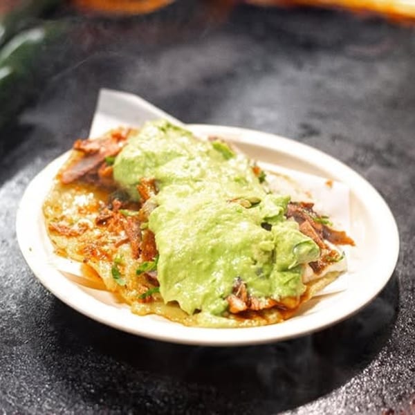 A Tacos El Franc taco with meat and guacamole on a white plate, on a dark surface.